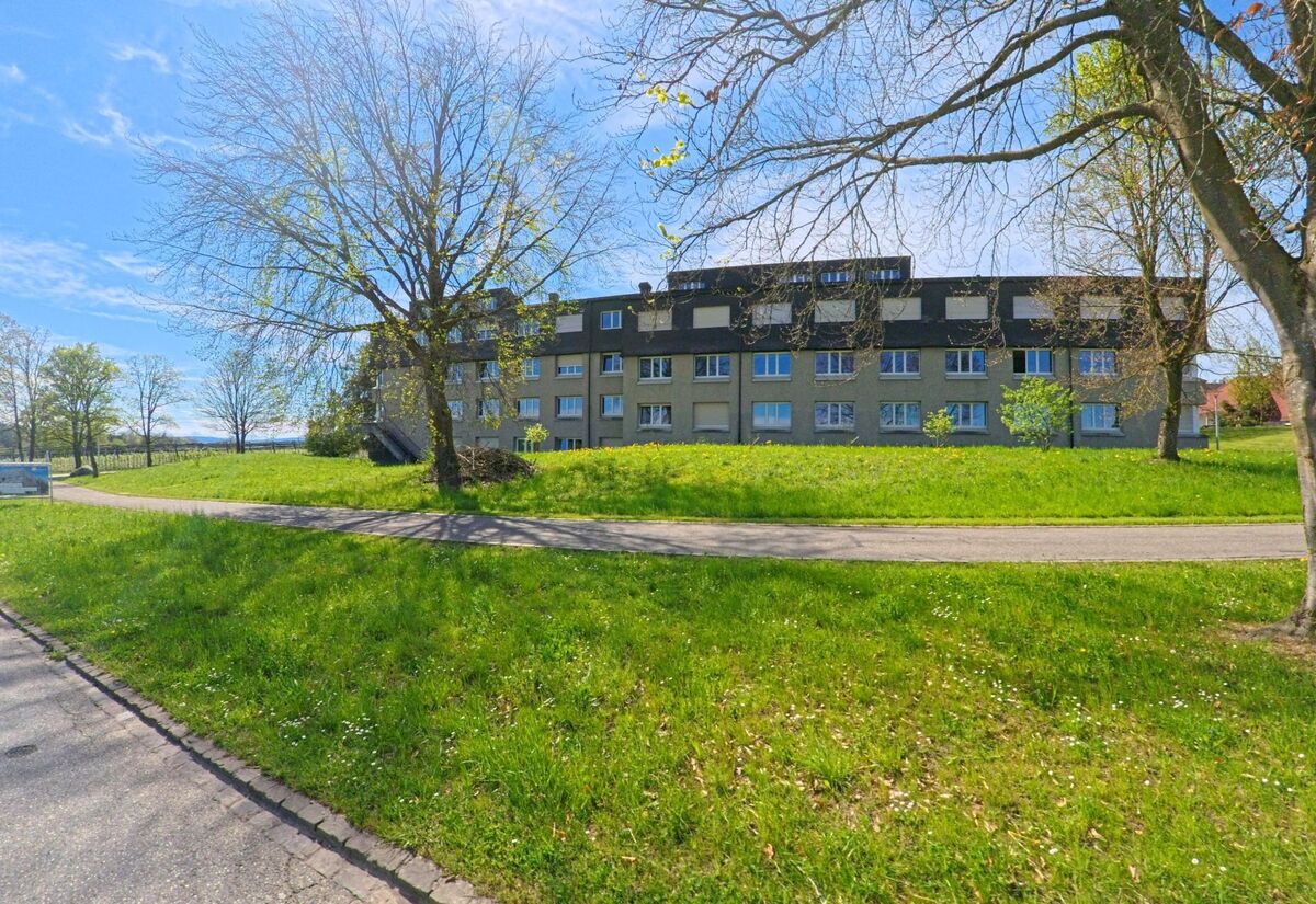 Vor einer mehrstöckigen Wohnanlage in Lindau (8315, Kanton Zürich) breitet sich eine gepflegte Grünfläche mit einzelnen Bäumen unter blauem Frühlingshimmel aus.