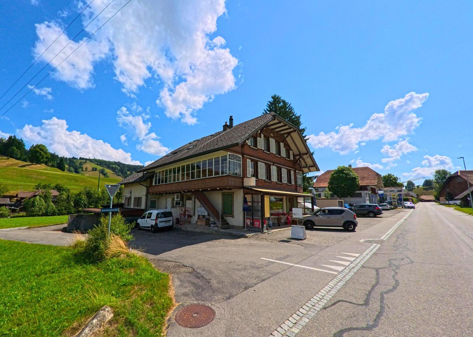 In Linden (3673, Kanton Bern) steht ein traditionelles Gebäude mit Laden im Erdgeschoss an einer verkehrsarmen Strasse, umgeben von grünen Hügeln und vereinzelten Bauernhäusern.