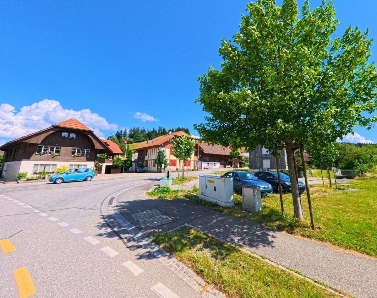 In einem Wohnquartier von Linden (3673, Kanton Bern) säumen traditionelle Häuser und geparkte Autos eine leicht gebogene Strasse mit Bäumen und Wiesen im Hintergrund.
