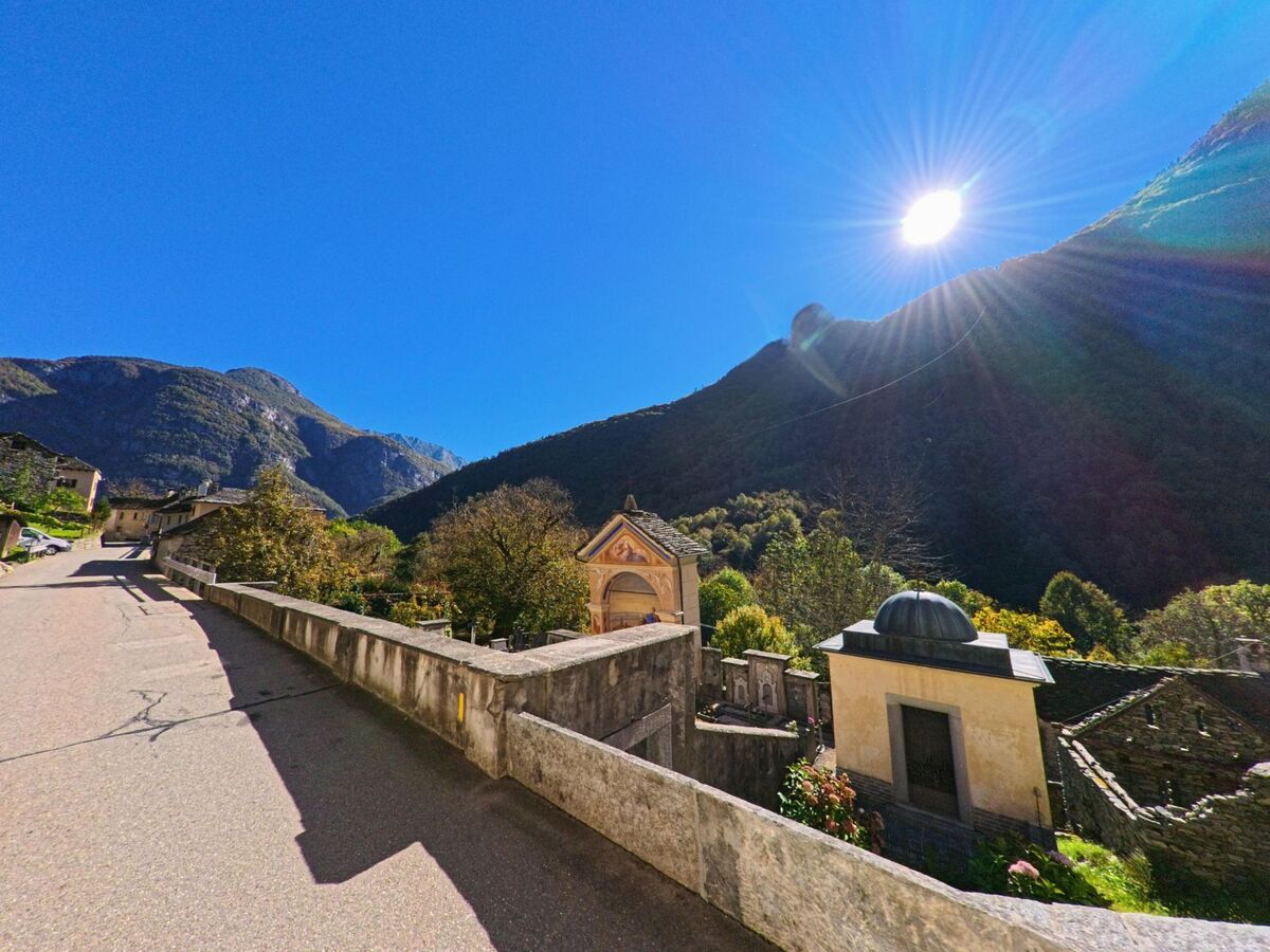 Bei strahlendem Sonnenschein führt in Linescio (6682, Kanton Tessin) eine Strasse an kleinen Kapellen und alten Steinhäusern vorbei, umgeben von bewaldeten Bergen.