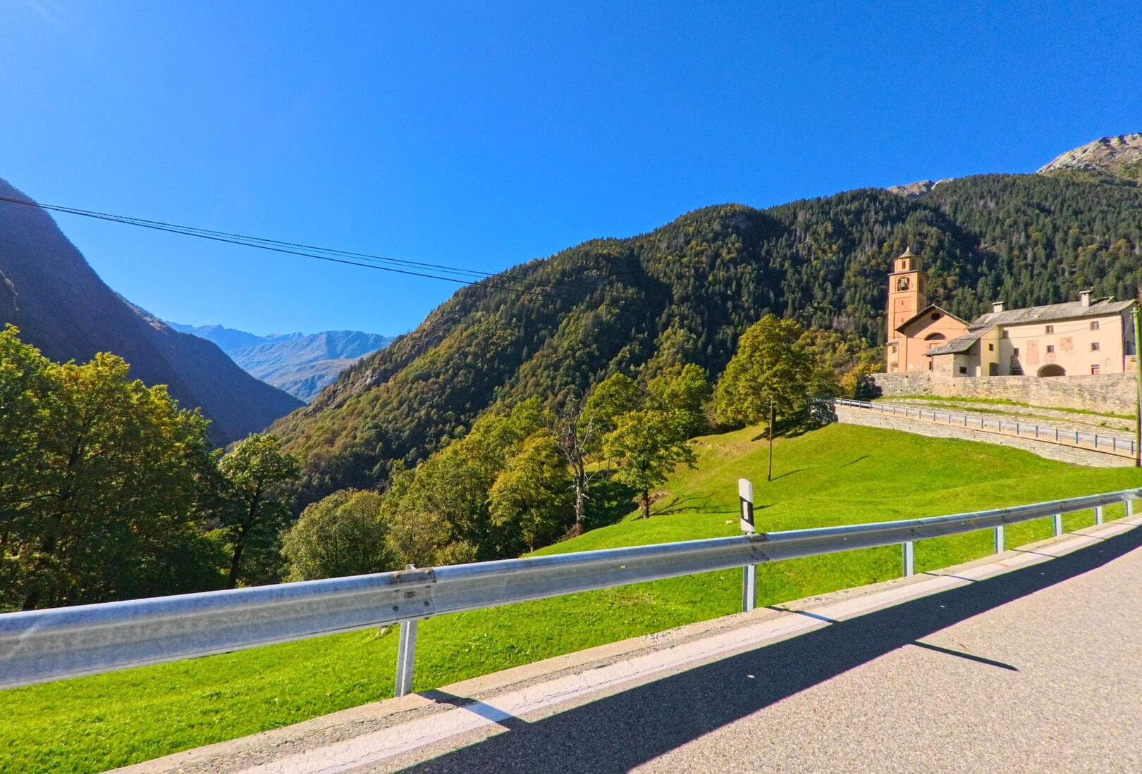 Die Kirche von Linescio (6682, Kanton Tessin) liegt malerisch am Hang über dem grünen Tal, umgeben von bewaldeten Bergen unter einem klaren blauen Himmel.
