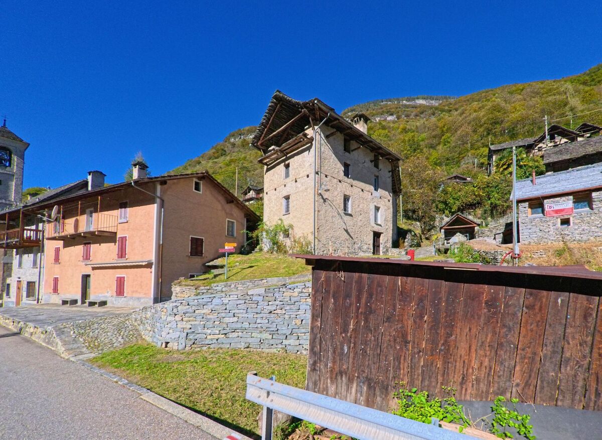 In Linescio (6682, Kanton Tessin) stehen historische Steinhäuser mit typischen Dächern aus Naturstein an einem sonnigen Hang mit Blick auf die umliegenden Berge.