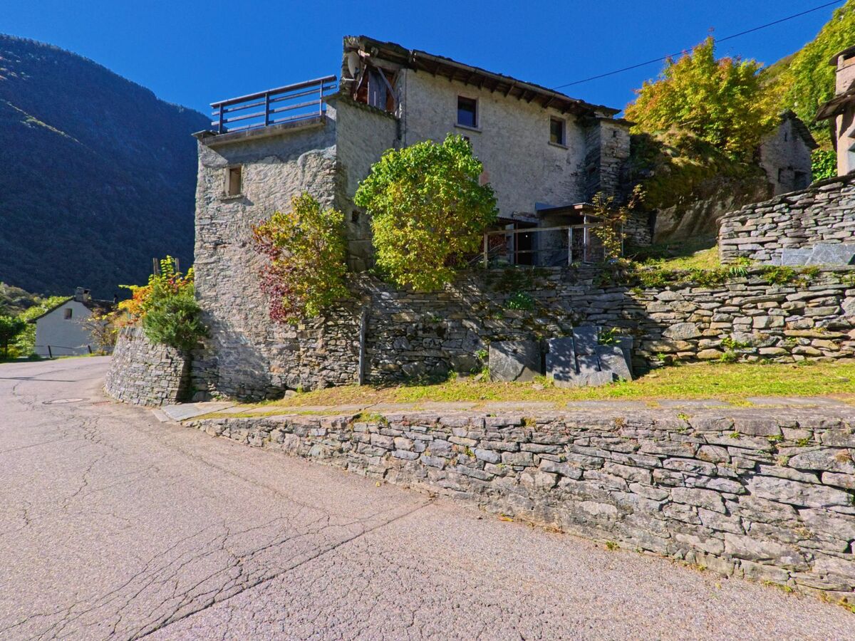 Ein traditionelles Steinhaus in Linescio (6682, Kanton Tessin) steht an einer Kurve der Dorfstrasse, umgeben von Natursteinmauern und herbstlich gefärbten Büschen.