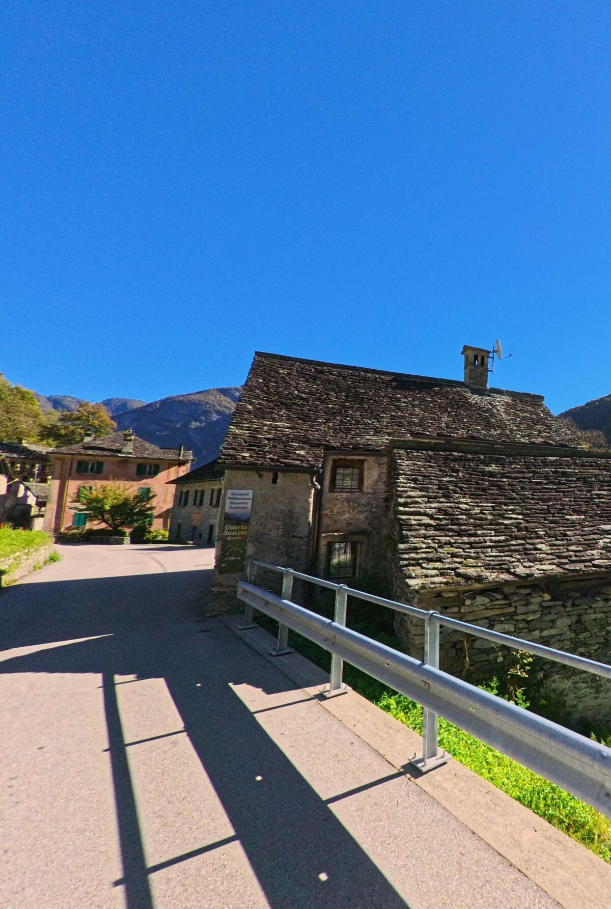 Ein altes Steinhaus mit Schieferdach in Linescio (6682, Kanton Tessin) steht an einer schmalen Strasse, die in das kleine Dorf mit weiteren historischen Gebäuden führt.
