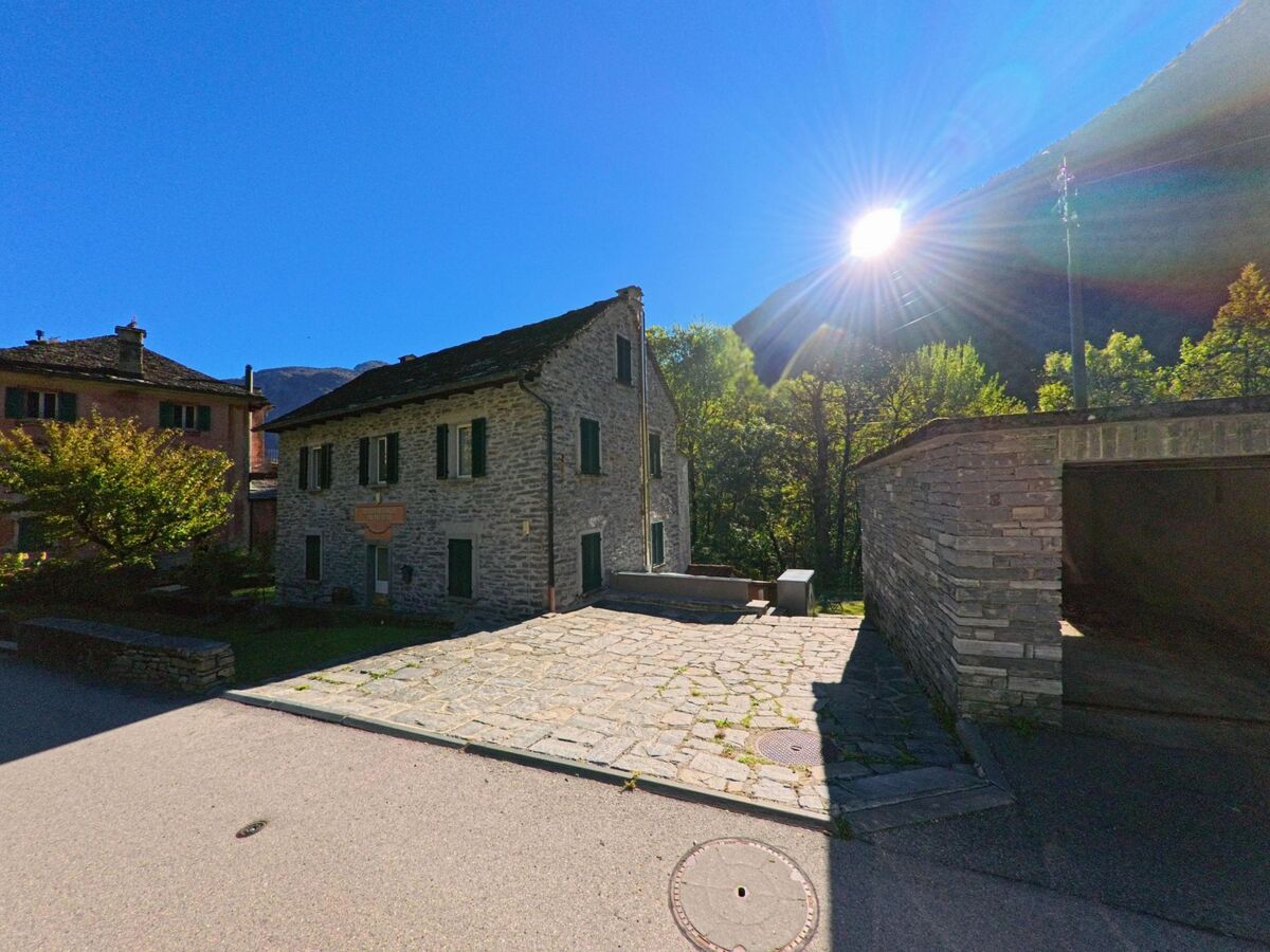 Ein helles Steinhaus in Linescio (6682, Kanton Tessin) steht am Rand einer kleinen gepflasterten Fläche, während die Sonne über den bewaldeten Hängen aufgeht.
