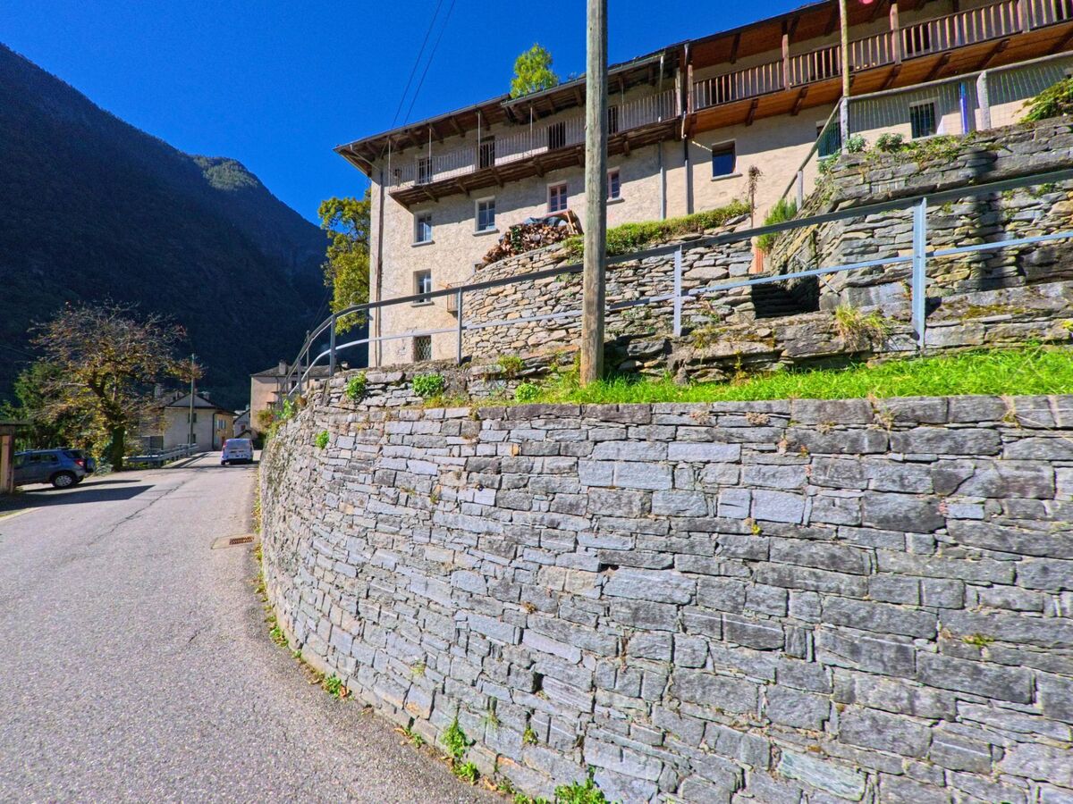Eine gepflasterte Strasse in Linescio (6682, Kanton Tessin) verläuft an einer Trockenmauer entlang, über der sich alte Wohnhäuser mit Holzbalkonen erheben.