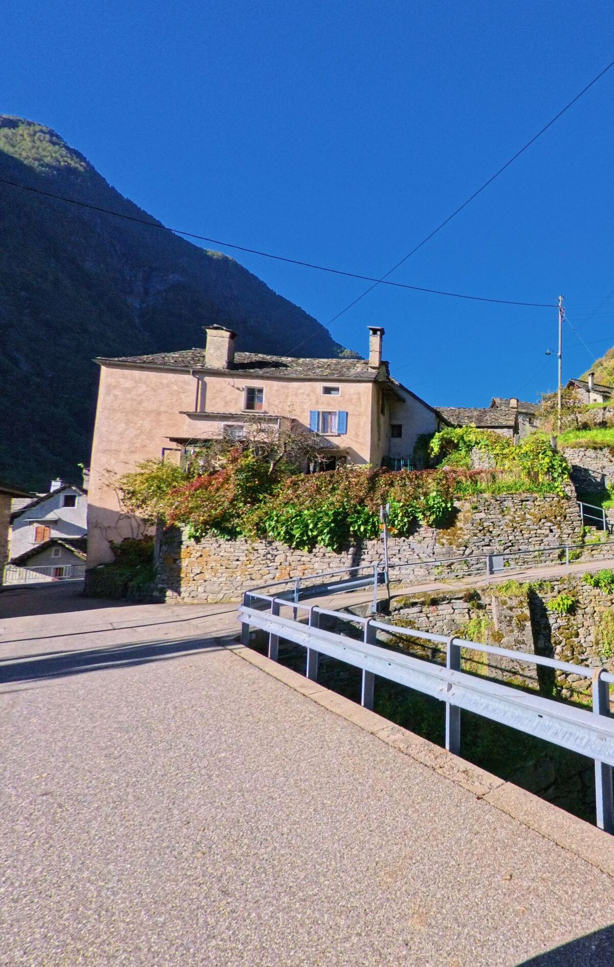Ein traditionelles Wohnhaus in Linescio (6682, Kanton Tessin) steht oberhalb einer gepflasterten Strasse, umgeben von Natursteinmauern und grünen Pflanzen im Sonnenschein.