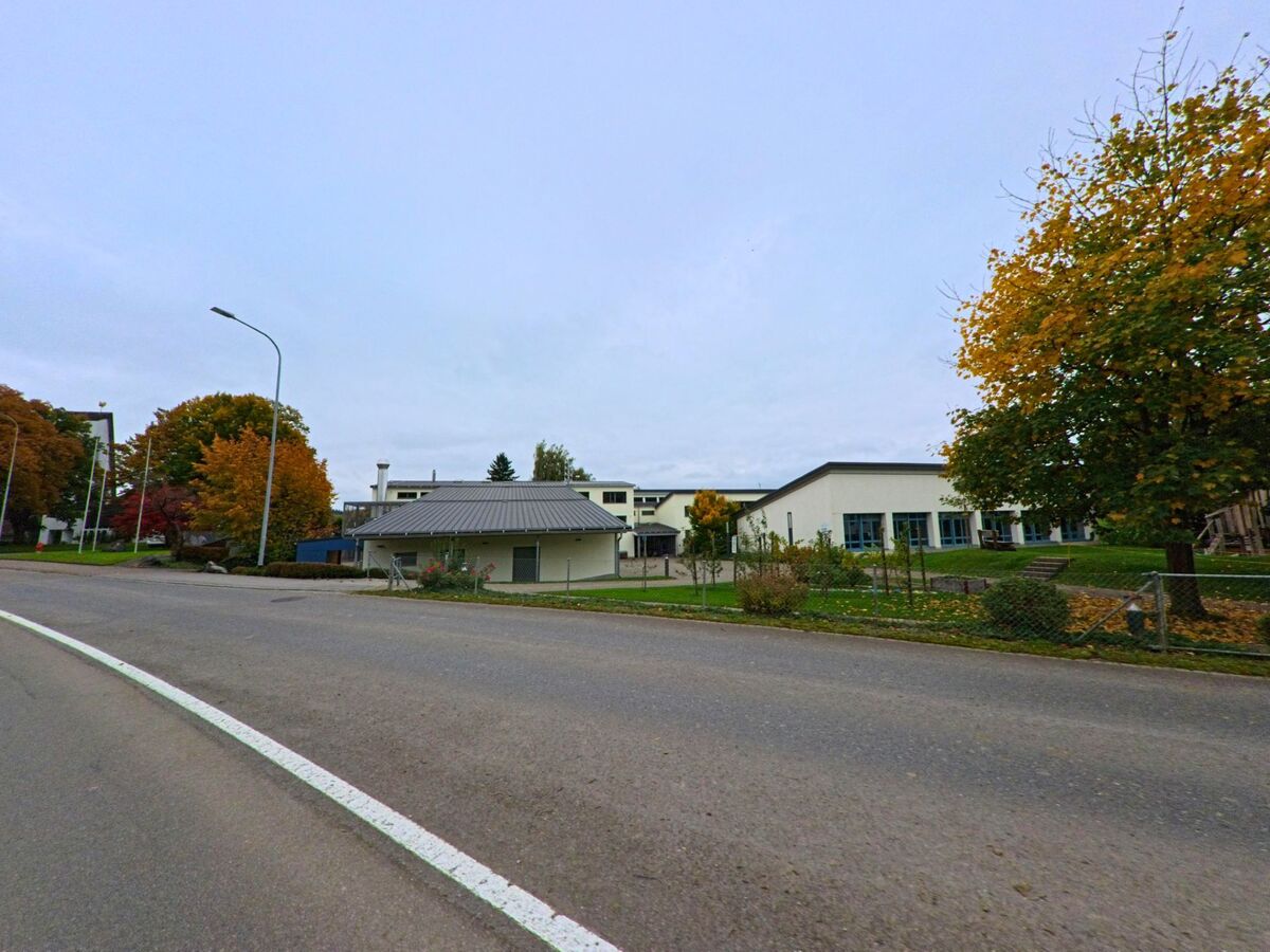 Hinter einer breiten Strasse in Lommis (9506, Kanton Thurgau) liegen flache Gebäude mit Grünflächen, eingerahmt von Bäumen in Herbstfarben.