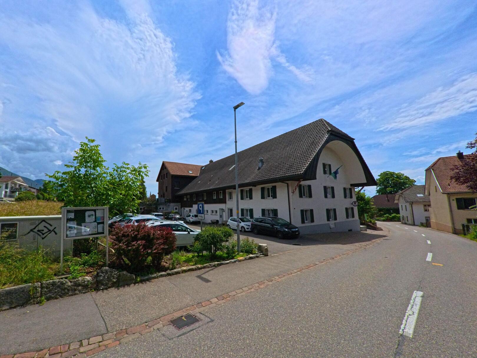 In Lommiswil (4514, Kanton Solothurn) führt eine schmale Strasse am Gemeindehaus mit markantem Rundgiebel und angrenzendem Parkplatz vorbei.