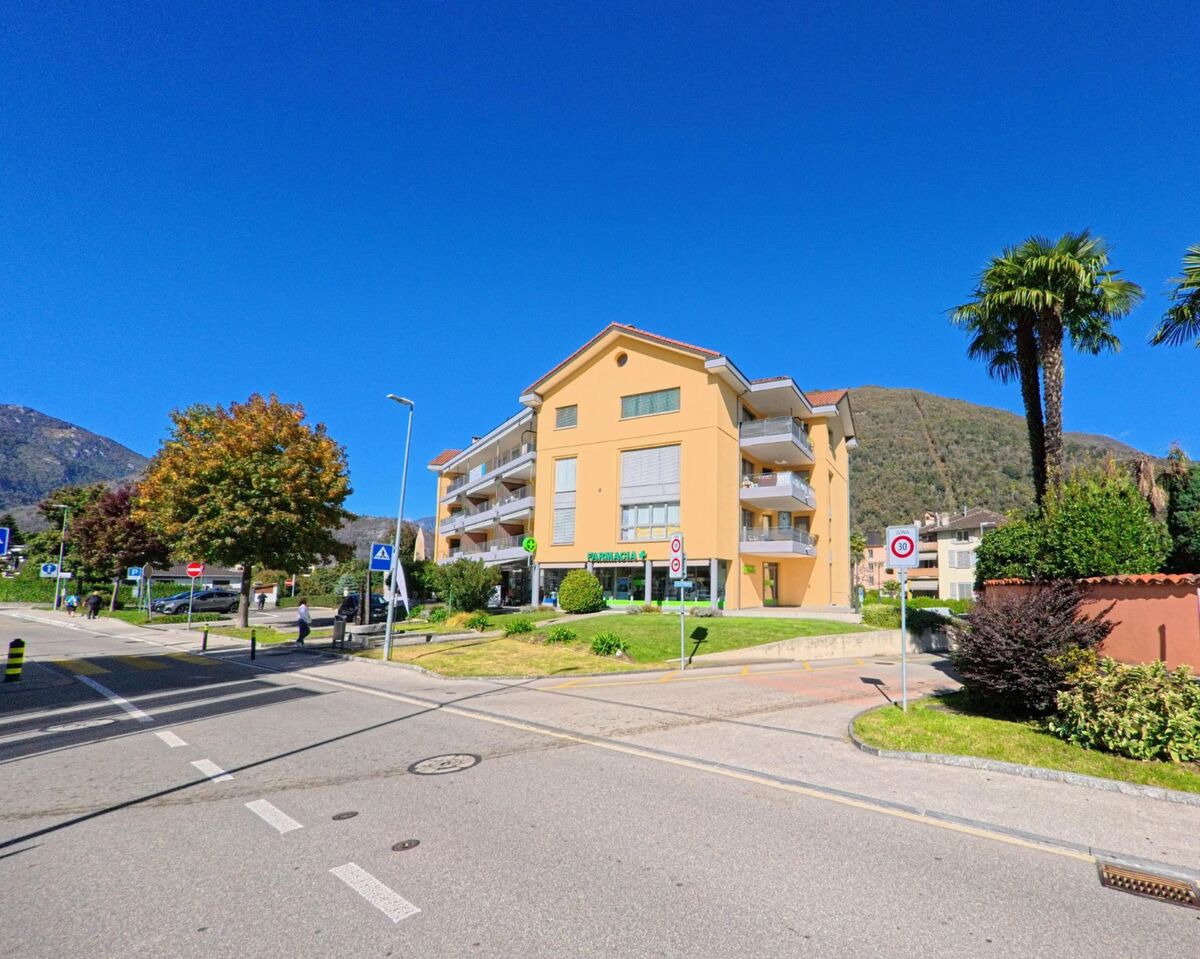 Ein modernes Wohnhaus mit Balkonen steht in Losone (6616, Kanton Tessin) an einer sonnigen Strasse mit Palmen und gepflegten Anlagen.