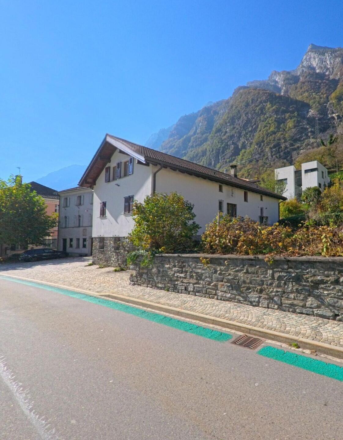 Entlang einer ruhigen Strasse verläuft in Lostallo (6558, Kanton Graubünden) ein Wohnhaus mit Natursteinmauer vor steil ansteigenden Berghängen.