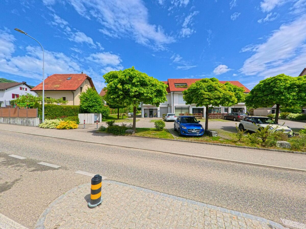 In Lostorf (4654, Kanton Solothurn) liegt ein Wohn- und Geschäftshaus mit rotem Ziegeldach hinter einem Parkplatz, der von gleichmässig geschnittenen Bäumen gesäumt ist.