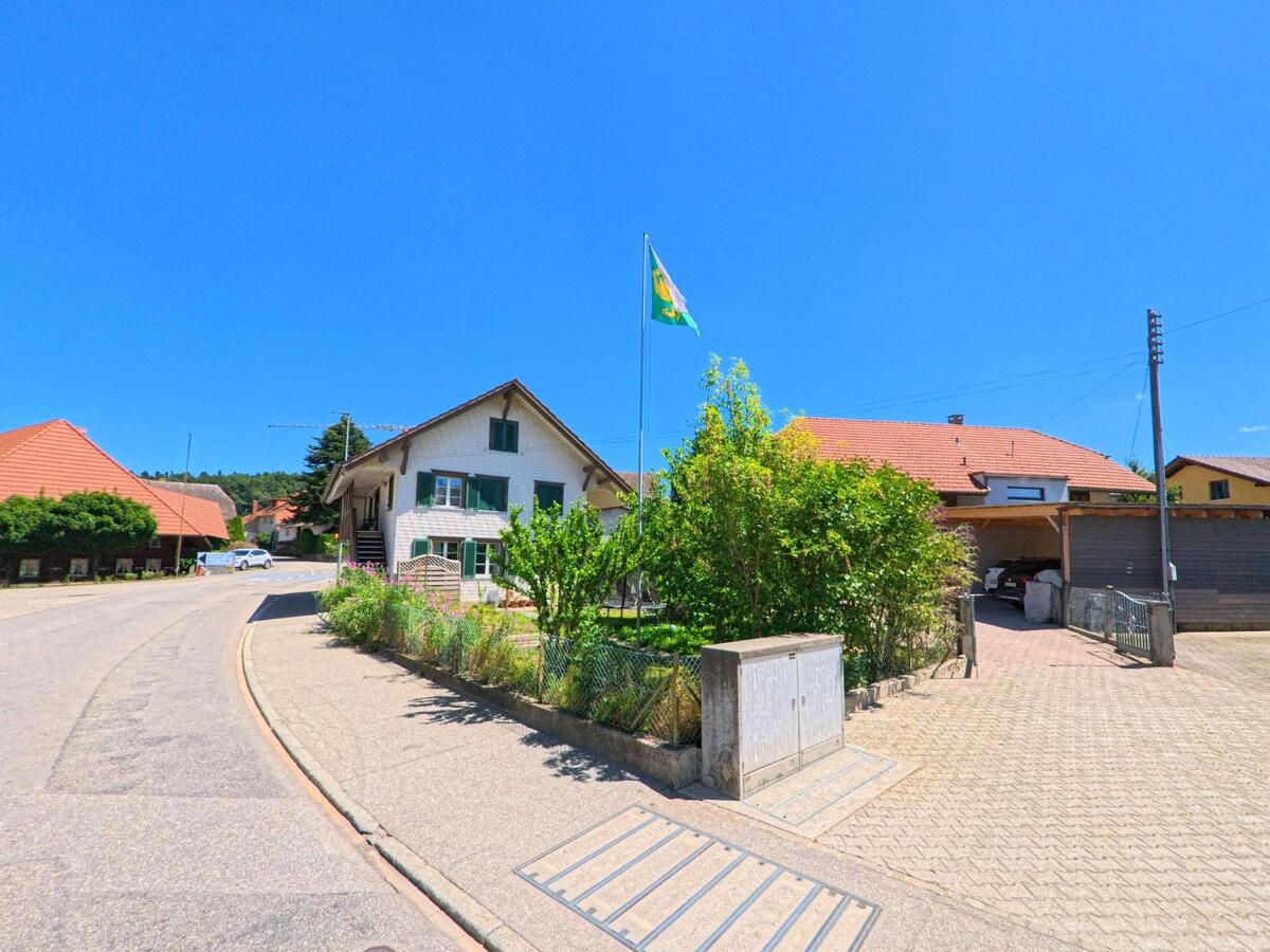An einer leicht ansteigenden Strasse in Lotzwil (4932, Kanton Bern) steht ein Haus mit gruener Fensterlaeden und Garten, darueber weht eine Fahne mit Gemeindewappen.
