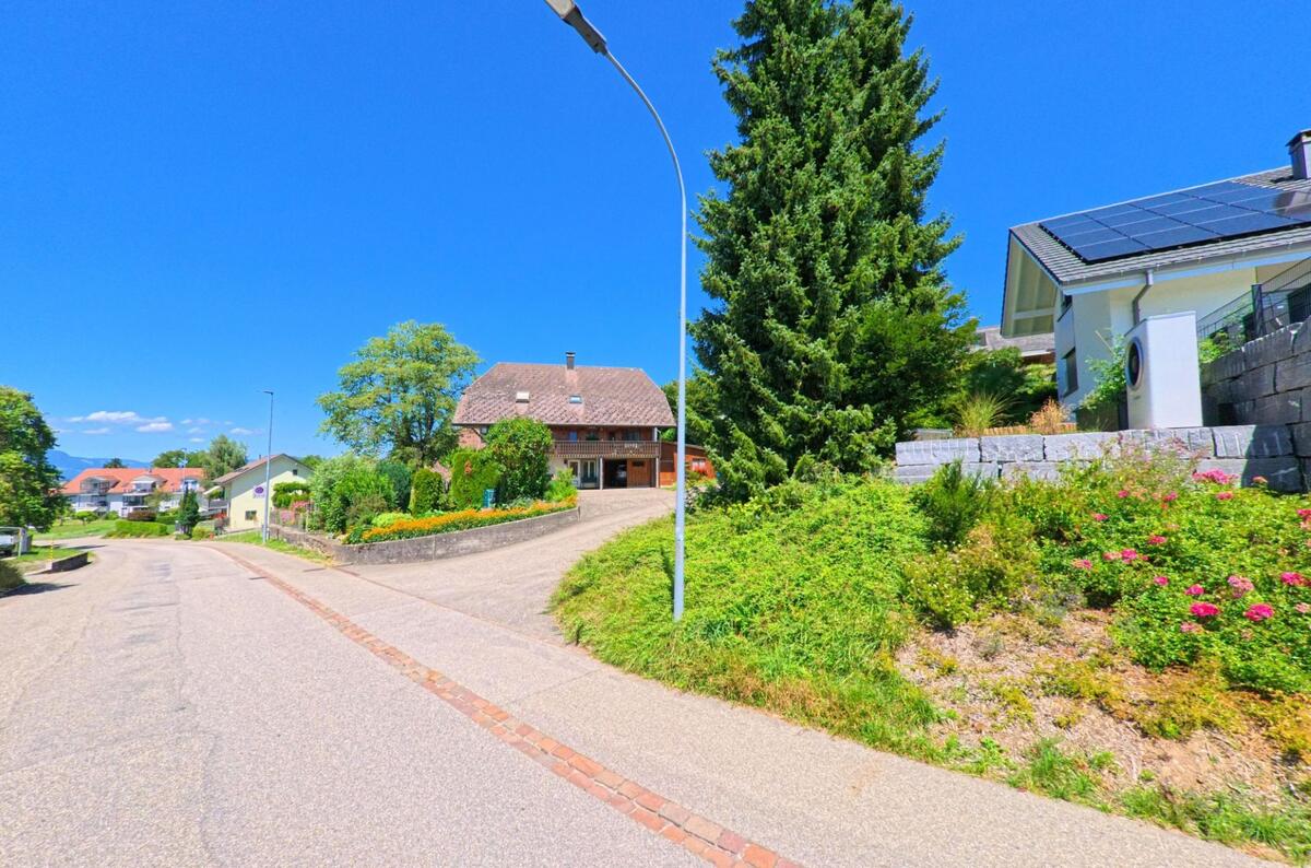 In Lotzwil (4932, Kanton Bern) fuehrt eine leicht ansteigende Strasse an gepflegten Gaerten, einem traditionellen Haus und modernen Wohnbauten mit Solarpanels vorbei.