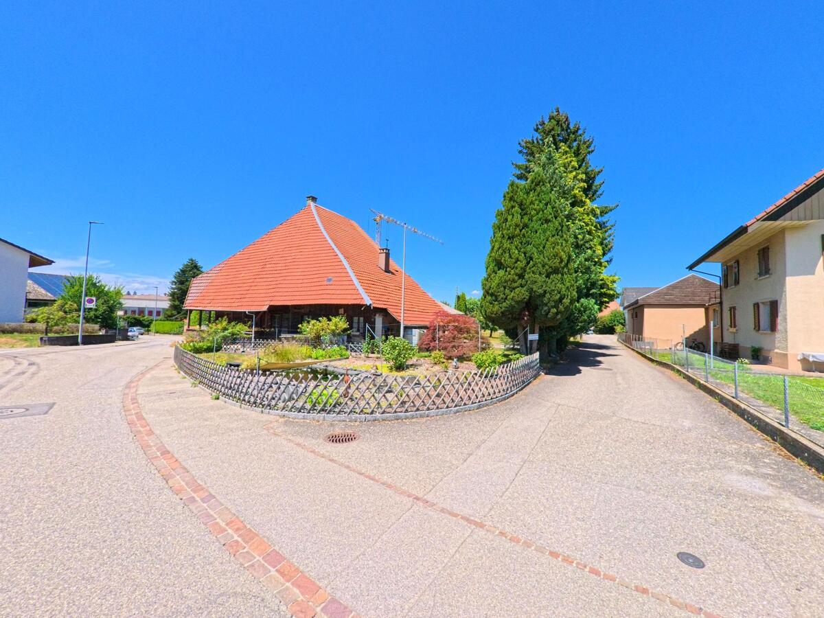 Ein auffaelliges Haus mit tief gezogenem Ziegeldach steht in Lotzwil (4932, Kanton Bern) an einer Strassengabelung, umgeben von gepflegten Gaerten und Gehwegen.
