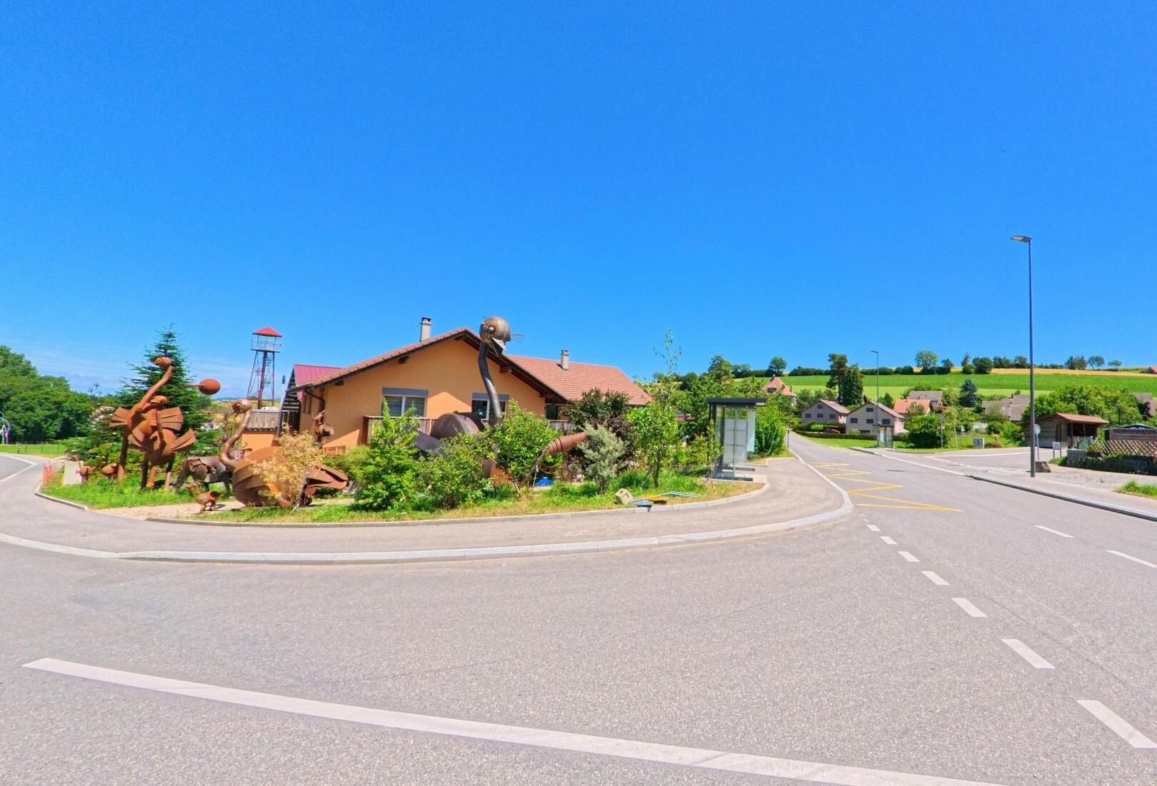 In Lucens (1683, Kanton Waadt) liegt ein kleines Wohnhaus mit originellen Metallskulpturen an einem Strassenkreisel, umgeben von Feldern und einzelnen Gebäuden.