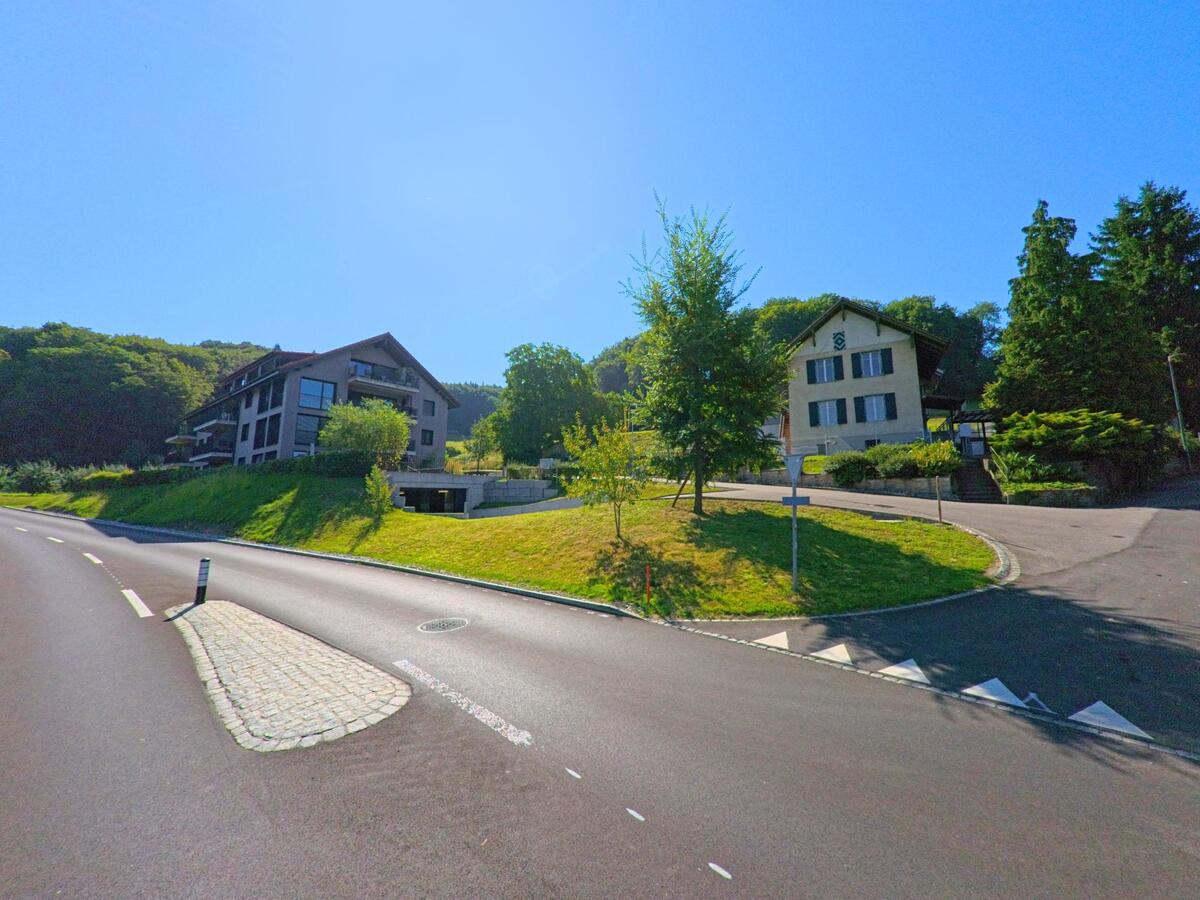 Am Rand von Lüscherz (2576, Kanton Bern) stehen moderne Wohngebäude und ein traditionelles Haus oberhalb einer breiten Strasse.