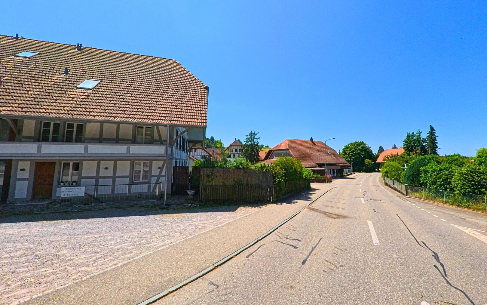 In Lüterkofen-Ichertswil (4571, Kanton Solothurn) verläuft eine ruhige Strasse mit traditionellen Wohnhäusern und gepflegten Gärten unter strahlend blauem Himmel.