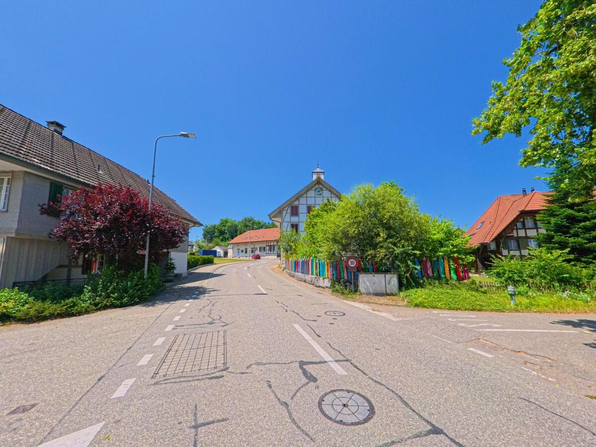 Eine leicht ansteigende Strasse in Lüterkofen-Ichertswil (4571, Kanton Solothurn) führt vorbei an traditionellen Häusern und einem bunt geschmückten Garten mit Sicht auf ein grosses Fachwerkhaus.
