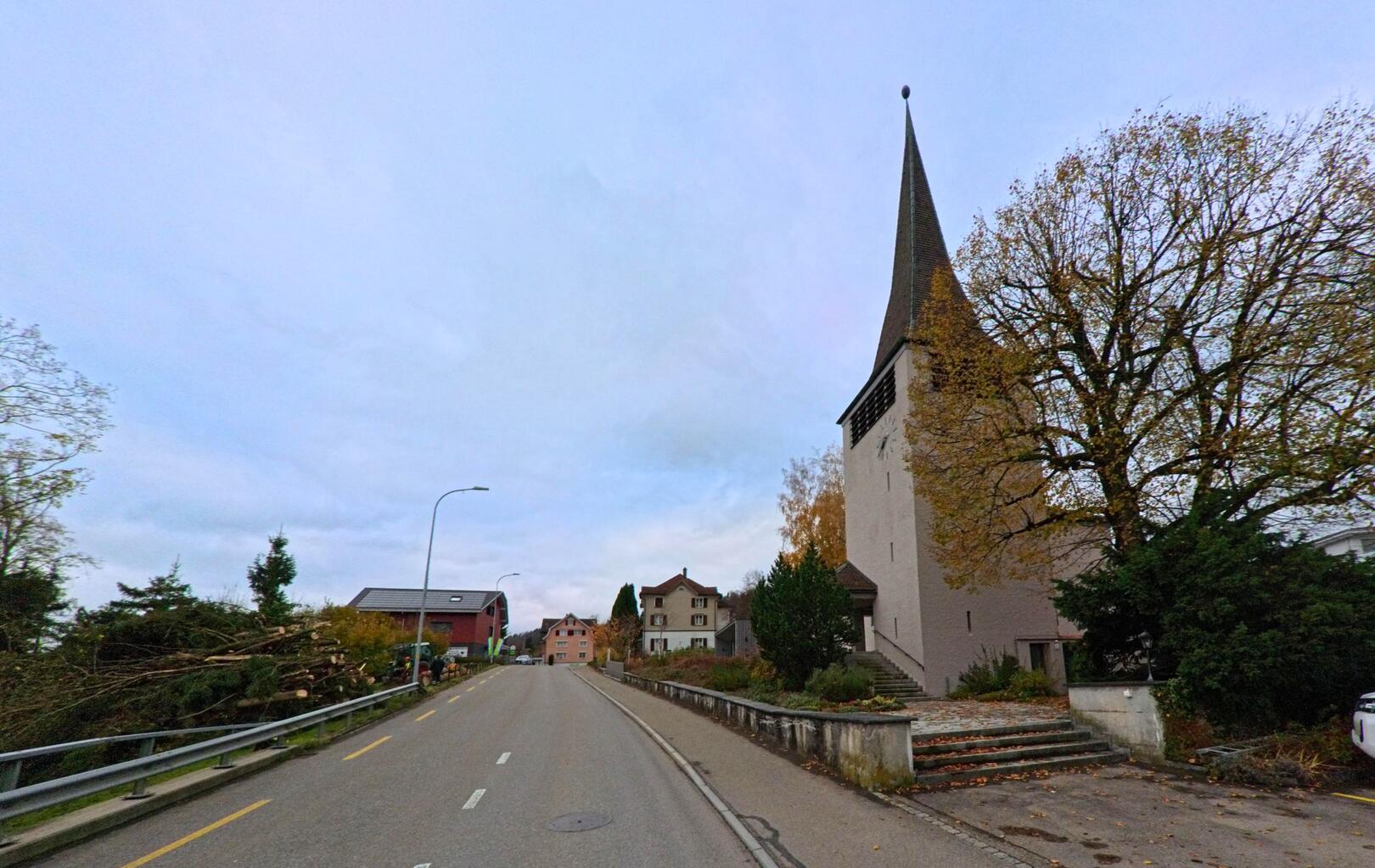 An der Hauptstrasse von Lütisburg (9604, Kanton St. Gallen) steht rechts ein Kirchturm neben herbstlichem Laub, waehrend sich die Bebauung zur Ortsmitte verdichtet.