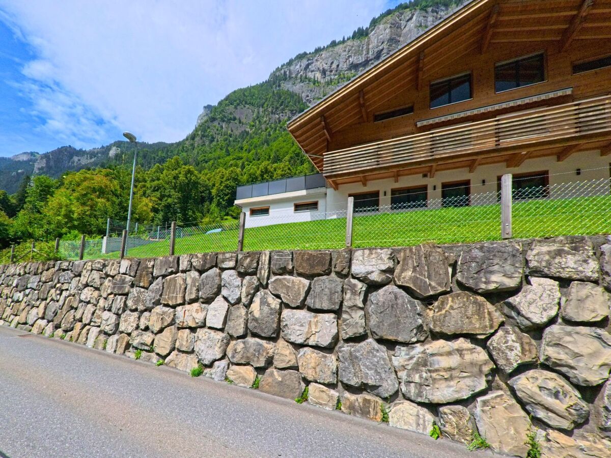 Ein modernes Haus mit Holzfassade und grossem Balkon erhebt sich in Lütschental (3816, Kanton Bern) über einer hohen Stützmauer aus Natursteinen vor steilen Berghängen.