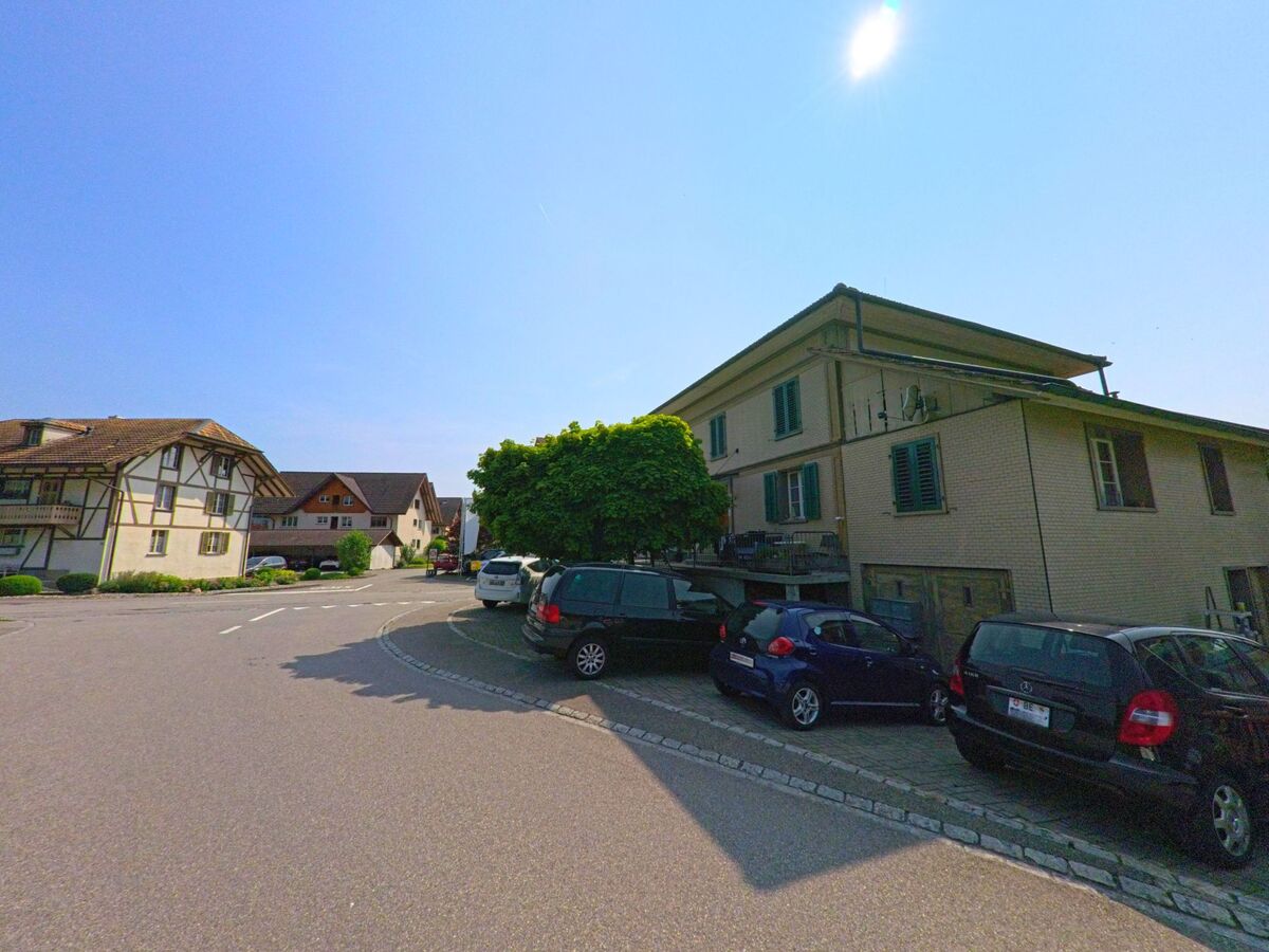  In Lützelflüh (3415, Kanton Bern) verläuft eine Wohnstrasse mit parkenden Autos entlang traditioneller und moderner Häuser unter klarem Himmel.