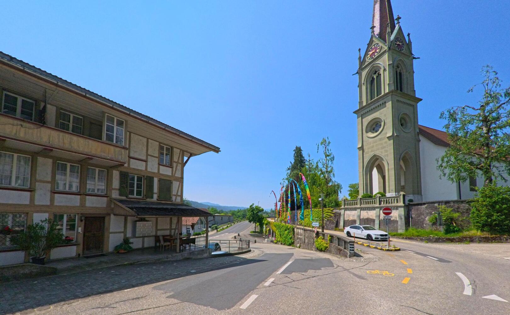 In Lützelflüh (3432, Kanton Bern) markiert der hohe Kirchturm mit Uhr den Übergang von einem historischen Fachwerkhaus zur offenen Landstrasse mit Radweg.