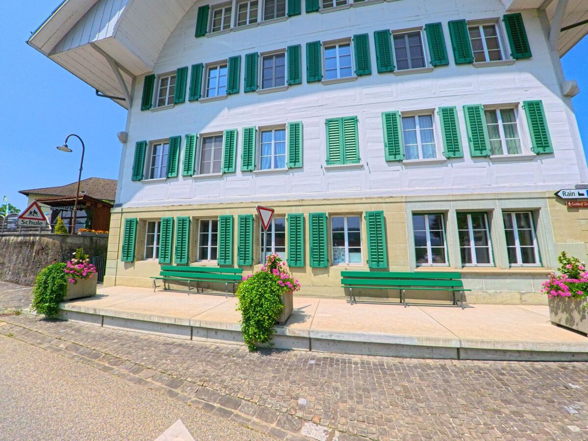 Vor einem Schulgebäude mit grünen Fensterläden in Lützelflüh (3432, Kanton Bern) laden Bänke und Blumenkübel zum Verweilen ein.