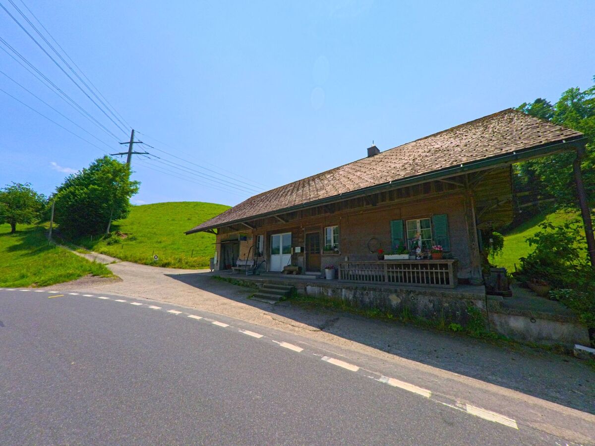  Ein traditionelles Bauernhaus mit gedeckter Veranda steht in Lützelflüh (3456, Kanton Bern) am Strassenrand, umgeben von grünen Hügeln und einem Kiesweg.