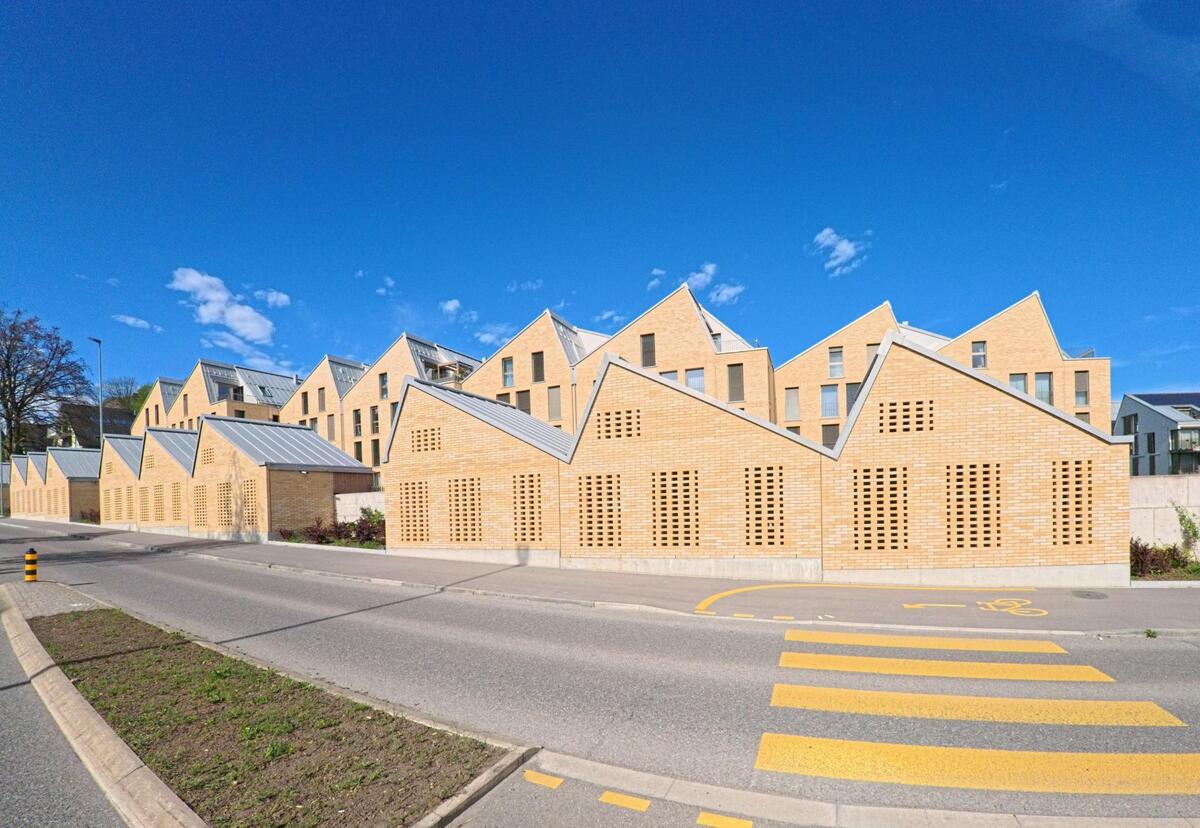 In Lufingen im Kanton Zürich, PLZ 8426, steht eine moderne Wohnanlage mit markanten Satteldächern und gelochten Backsteinfassaden direkt an einer Quartierstrasse mit Zebrastreifen.