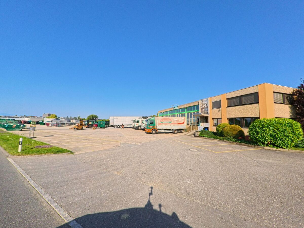 Auf einem grossen Firmengelaende in Lully (1470, Kanton Freiburg) stehen mehrere Lastwagen und Container vor einem Industriegebaeude bei klarem Himmel.