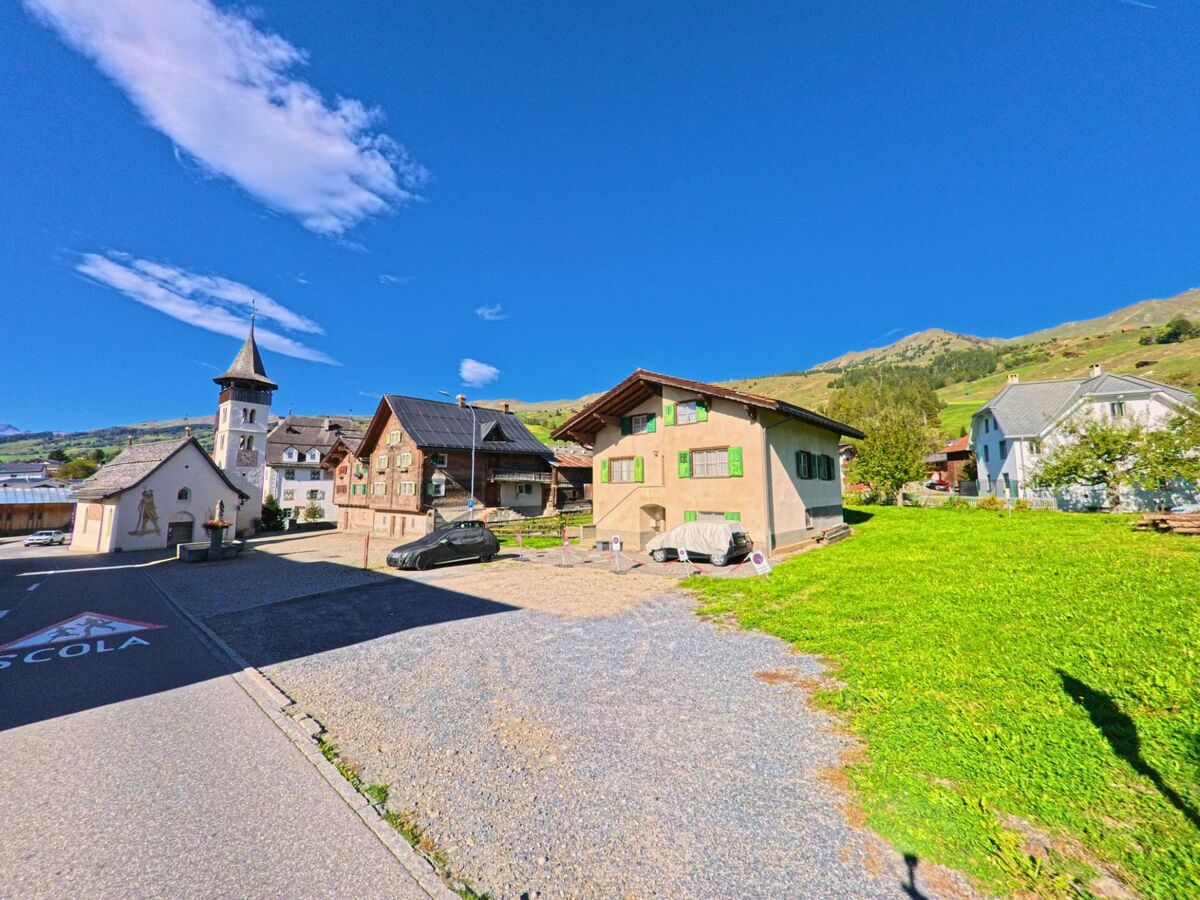 Im Dorfkern von Lumnezia (7143, Kanton Graubuenden) steht eine kleine Kirche mit Spitzdach neben traditionellen Wohnhaeusern und Wiesen unter klarem Himmel.