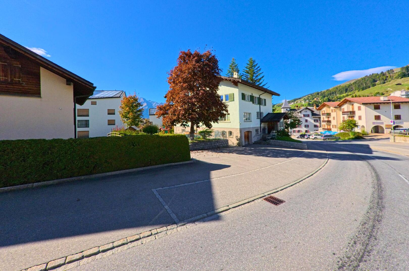 An einer leicht gekruemmten Strasse in Lumnezia (7145, Kanton Graubuenden) stehen gepflegte Wohnhaeuser und ein Baum mit rotem Laub vor dem Hintergrund der Berge.