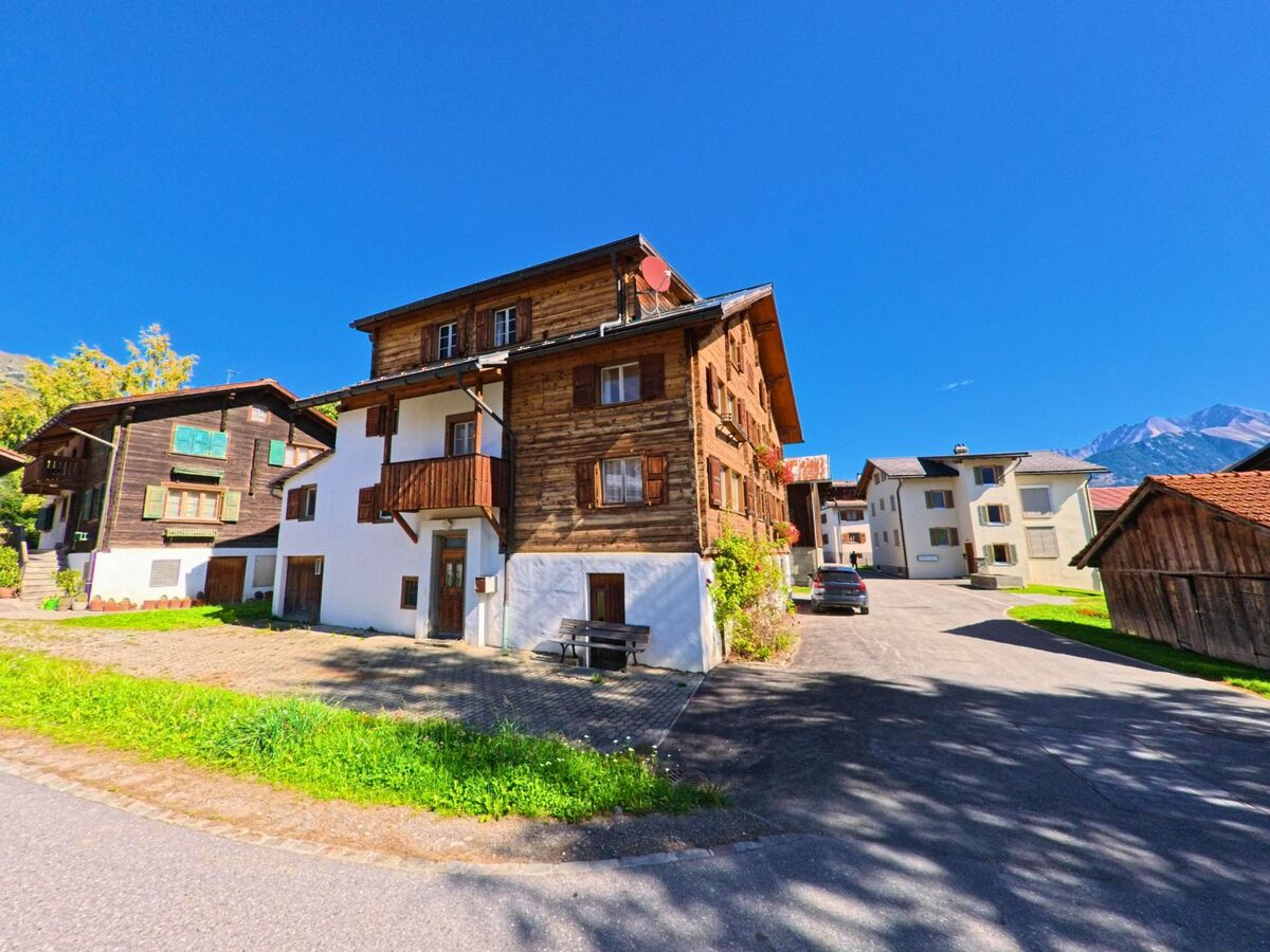 Traditionelle Holzhäuser mit Balkonen und Fensterläden praegen in Lumnezia (7145, Kanton Graubuenden) das Dorfbild unter blauem Himmel und Bergkulisse.