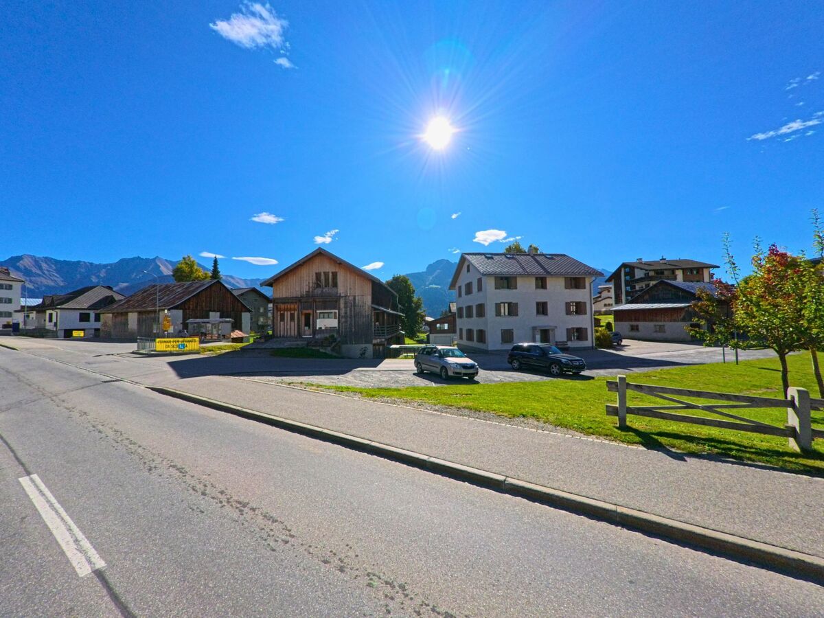 In Lumnezia (7145, Kanton Graubuenden) stehen mehrere Wohn- und Holzhaeuser an einer sonnigen Strasse mit Blick auf die umliegenden Berge.