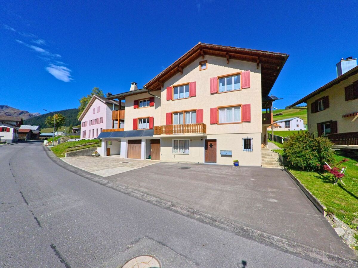 An einer ruhigen Strasse in Lumnezia (7148, Kanton Graubuenden) stehen helle Wohnhaeuser mit roten Fensterlaeden vor einer gruenen Hügellandschaft.