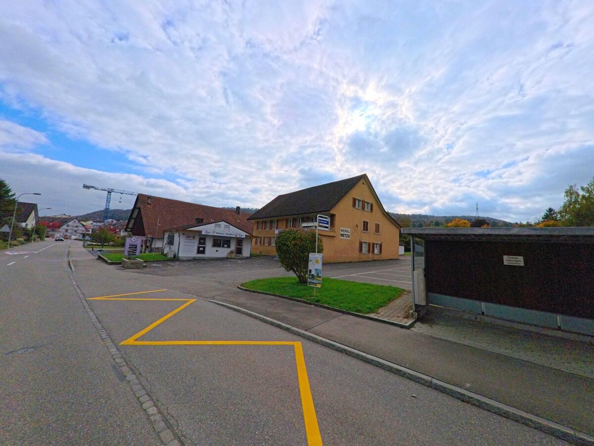 Entlang der Hauptstrasse in Lupfig (5242, Kanton Aargau) stehen Gewerbegebäude und Wohnhäuser, im Hintergrund ist ein Baukran über dem Dorf zu sehen.
