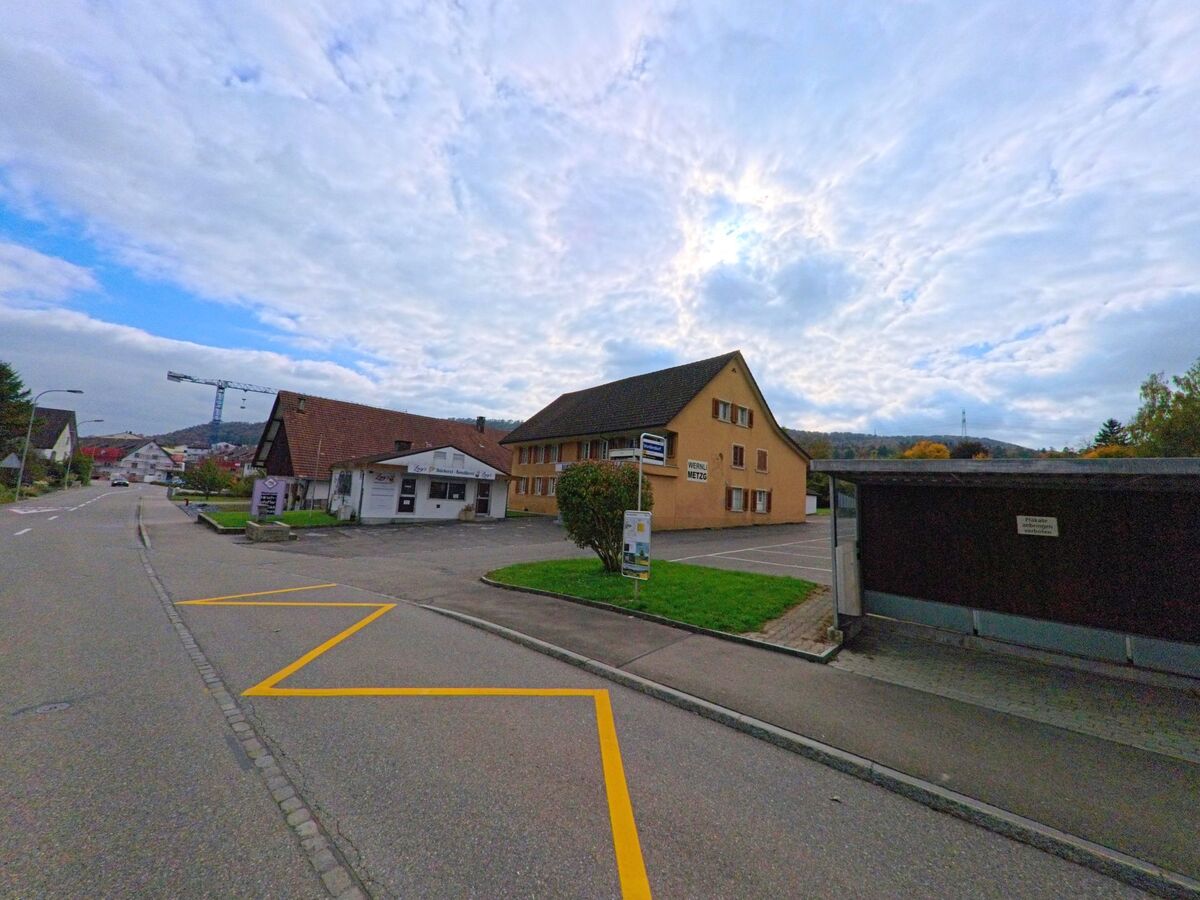 Entlang der Hauptstrasse in Lupfig (5242, Kanton Aargau) stehen Gewerbegebäude und Wohnhäuser, im Hintergrund ist ein Baukran über dem Dorf zu sehen.