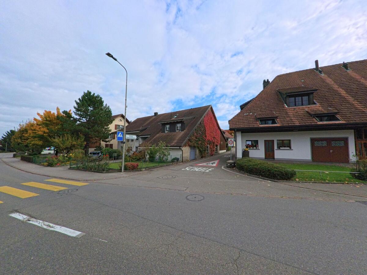 An einer Strassenkreuzung in Lupfig (5242, Kanton Aargau) stehen Wohnhäuser mit gepflegten Vorgärten und eine Scheune, deren Fassade von rotem Efeu bedeckt ist.
