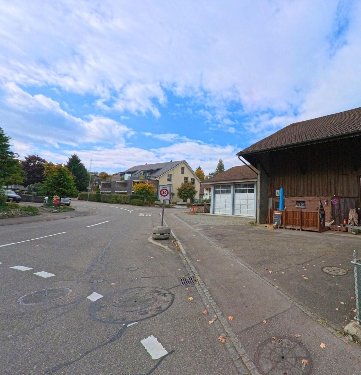 Eine Seitenstrasse in Lupfig (5242, Kanton Aargau) führt an einer alten Scheune und modernen Wohnhäusern vorbei, in einer verkehrsberuhigten Zone mit Tempo-30-Schild.