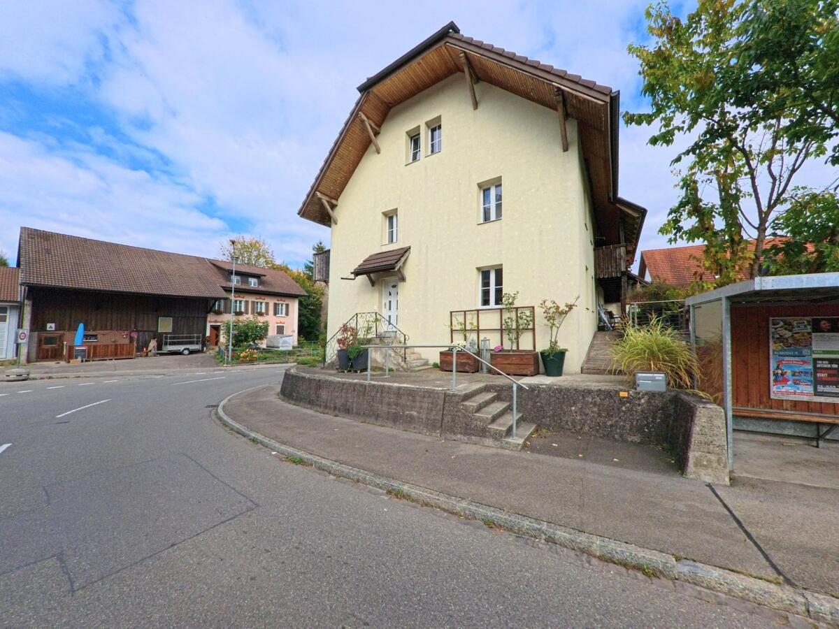 Ein helles Wohnhaus mit Treppe und Vorgarten steht an einer Strassenkurve in Lupfig (5242, Kanton Aargau), neben einer kleinen Bushaltestelle und älteren Gebäuden.