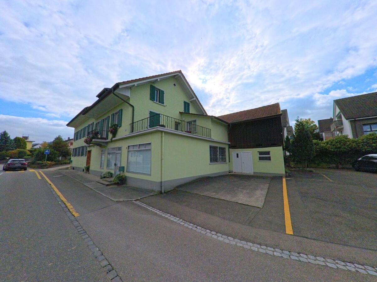 Ein hellgrünes Wohnhaus mit Fensterläden und Balkon steht an einer Strasse in Lupfig (5242, Kanton Aargau), neben einem kleinen Parkplatz und weiteren Einfamilienhäusern.
