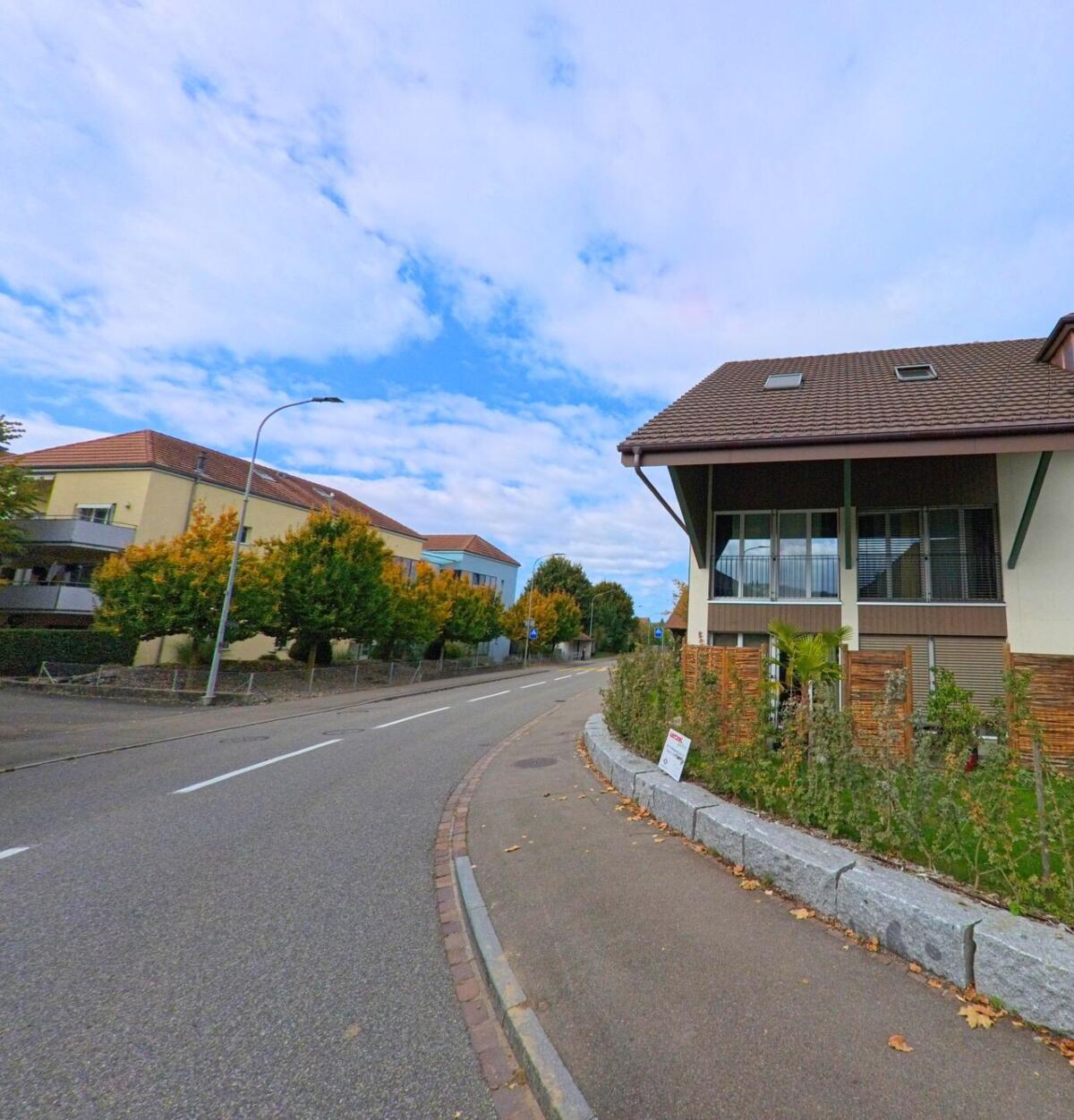 Entlang einer leicht gebogenen Strasse in Lupfig (5242, Kanton Aargau) stehen moderne Wohnhäuser mit Balkonen und herbstlich gefärbten Bäumen.