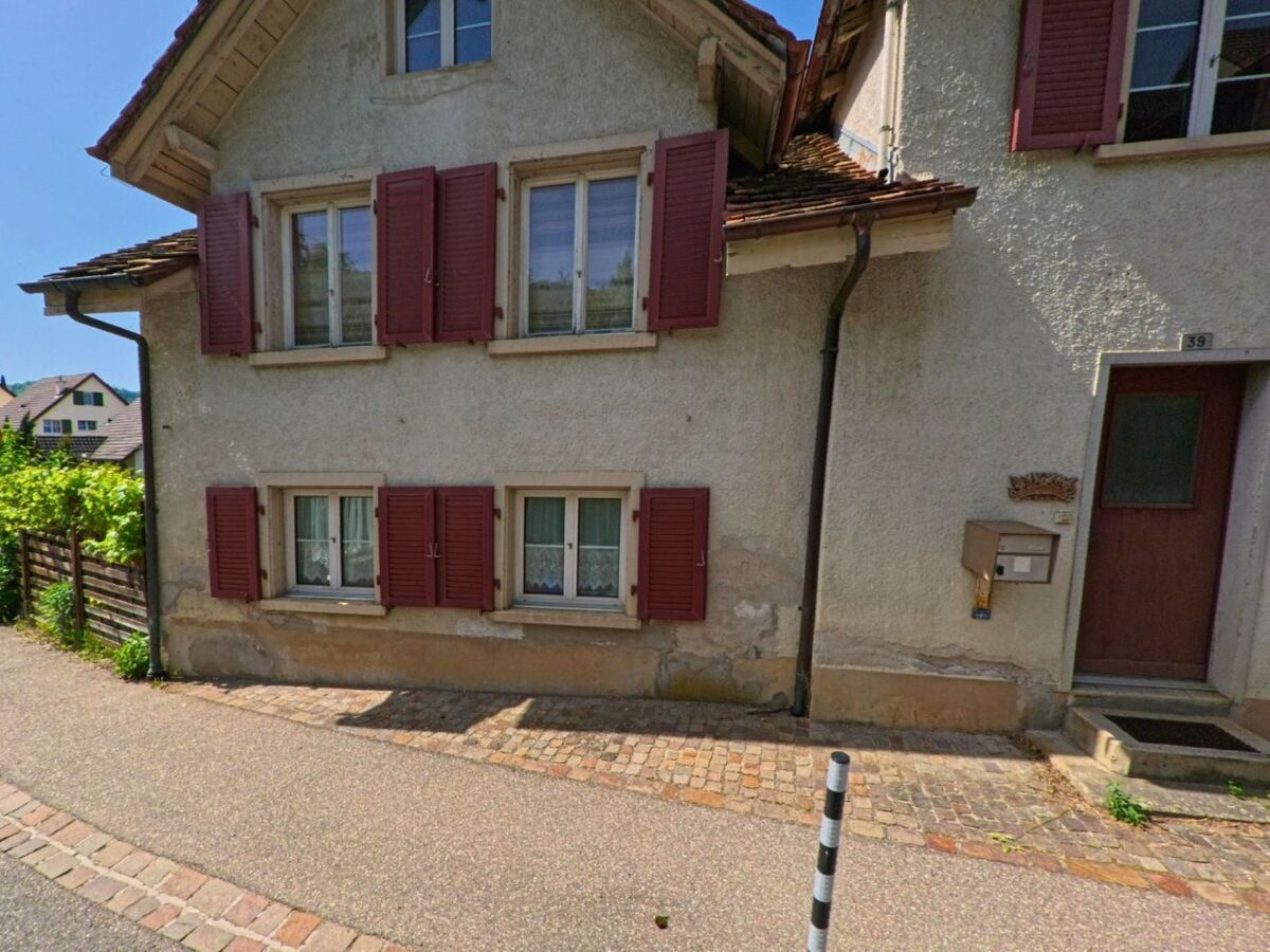 In Lupsingen (4419, Kanton Basel-Landschaft) steht ein älteres Wohnhaus mit roten Fensterläden direkt an einer schmalen, gepflasterten Gasse.