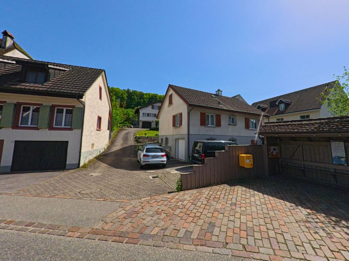 In Lupsingen (4419, Kanton Basel-Landschaft) führt eine gepflasterte Einfahrt zwischen Wohnhäusern hinauf zu weiteren Gebäuden am Waldrand, neben einem gelben Briefkasten.