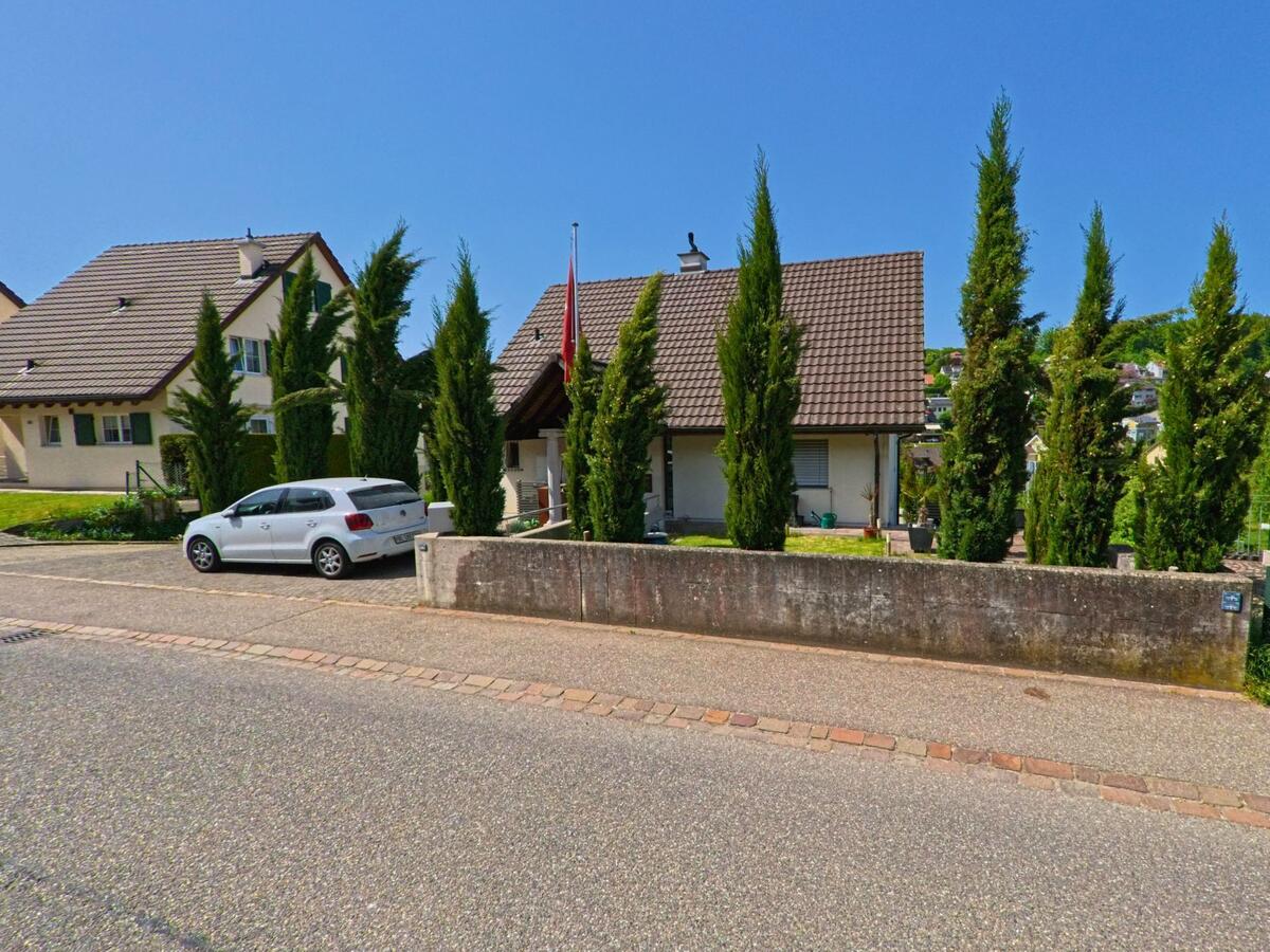 In Lupsingen (4419, Kanton Basel-Landschaft) steht ein Einfamilienhaus mit vorgelagerten Zypressen und Schweizer Flagge in einer ruhigen Wohnstrasse.