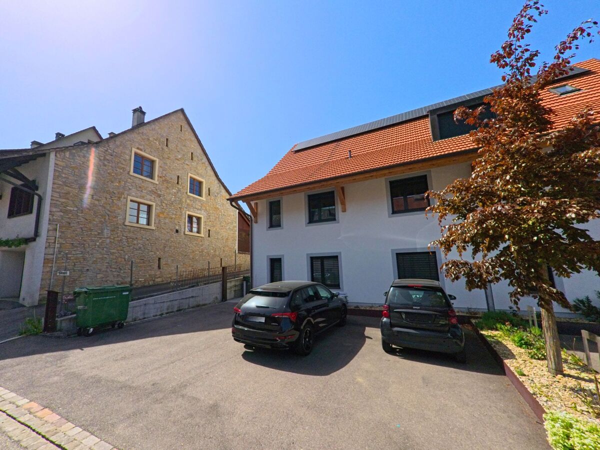 In Lupsingen (PLZ 4419, Kanton Basel-Landschaft) grenzen moderne Wohnhäuser mit roten Ziegeldächern an einen kleinen Parkplatz mit zwei Autos.