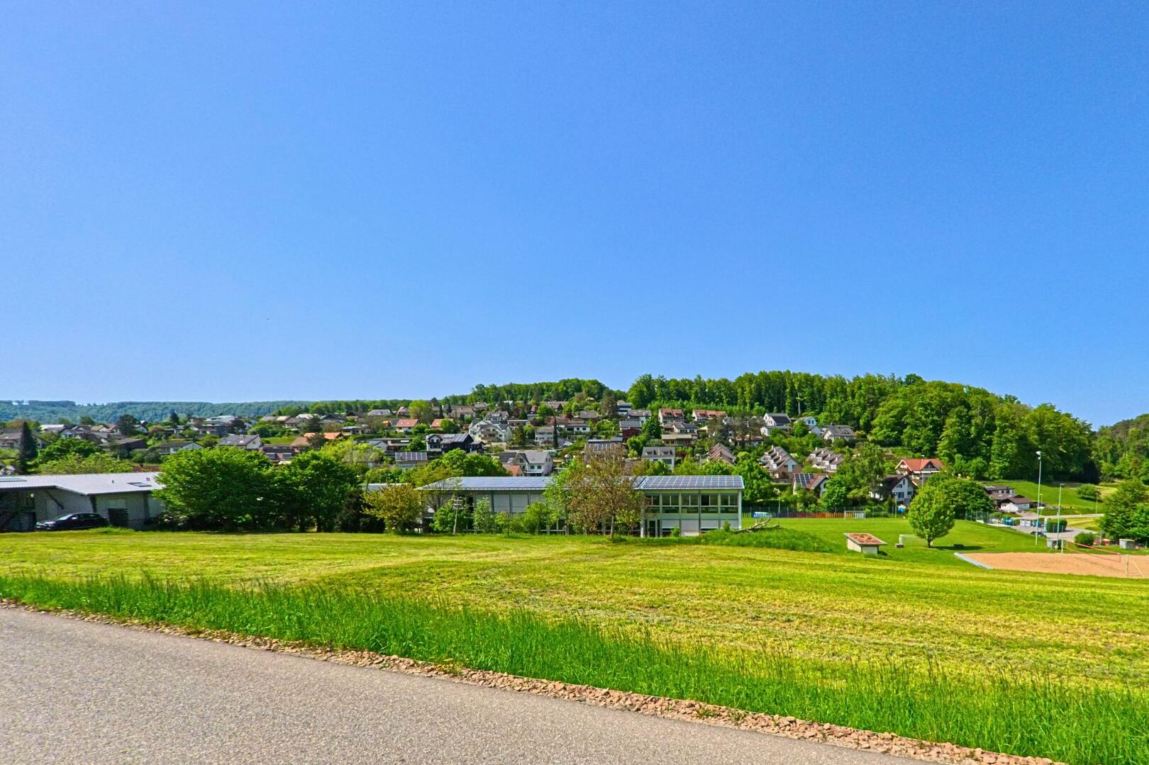 Von einem leicht erhöhten Punkt aus bietet sich ein weiter Blick über Lupsingen (4419, Kanton Baselland) mit seinen Wohnhäusern am Waldrand und den Sportanlagen im Vordergrund.