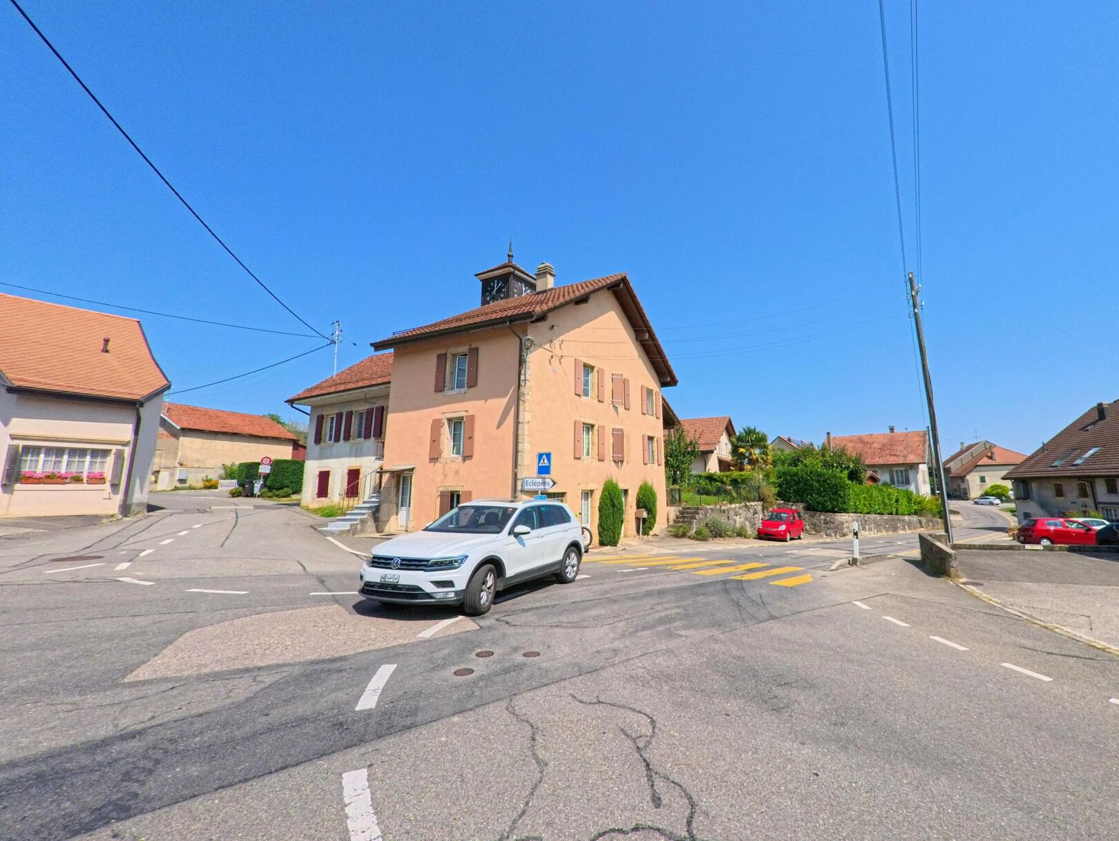 Im Zentrum von Lussery-Villars (1307, Kanton Waadt) steht ein Gebaeude mit Turmuhr an einer Strassenkreuzung, umgeben von weiteren Dorfhaeusern.
