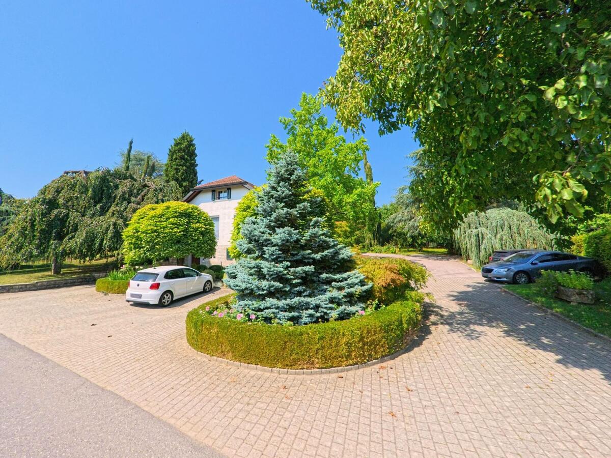 Ein gepflegter Garten mit rund geschnittenen Baeumen und einer zentralen Zierhecke umgibt ein Wohnhaus in Lussery-Villars (1307, Kanton Waadt).